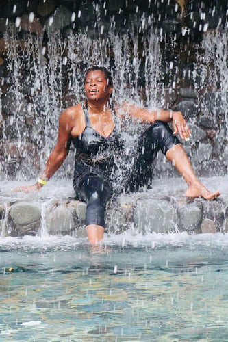 A woman of color sits under a waterfall at Ecotermales Hot Springs in Costa Rica, near Arenal Volcano. In a black swimsuit, she relaxes with one leg in the water, eyes closed in peaceful reflection.